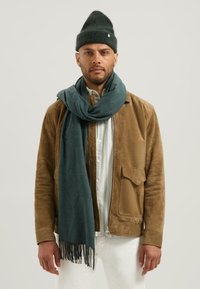 Hnědá semišová bunda přes světle kostkovanou košili, tmavě zelený vlněný šál s třásněmi a tmavě zelená pletená čepice.