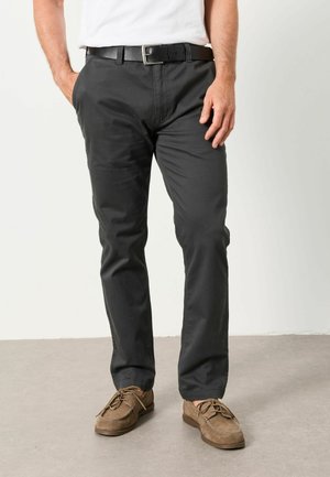 Mann trägt schwarze Hose mit geradem Bein, schwarzen Gürtel, braune Wildlederschnürschuhe und ein weißes Hemd mit den Händen in den Taschen.