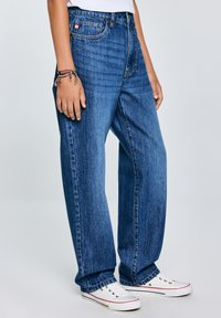 Jeans in denim azzurro chiaro con vita alta, design a gamba larga e stile classico a cinque tasche. Indossati con scarpe da ginnastica bianche.