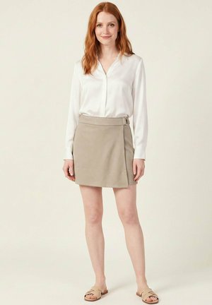 Femme aux cheveux roux portant un chemisier blanc, une jupe portefeuille beige avec un détail boucle et des sandales beige, debout devant un fond neutre.