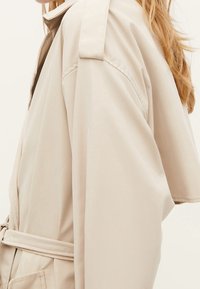 Beżowy trencz wykonany z gładkiej tkaniny, z paskiem w talii, szerokimi rękawami i pagonami na ramionach. Minimalistyczny design bez zdobień.