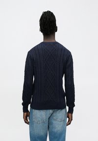 Person mit kurzen Dreadlocks, der einen marineblauen Kabelstrickpullover und hellblaue Jeans trägt und mit dem Rücken zu sehen ist, vor einem schlichten weißen Hintergrund.
