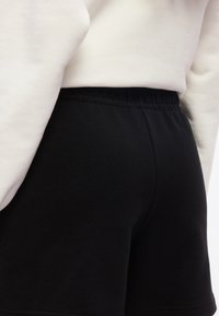 Κοντινό πλάνο ατόμου που φοράει μαύρο σορτς με ελαστική μέση και λευκό μακρυμάνικο μπλουζάκι σε απλό φόντο.