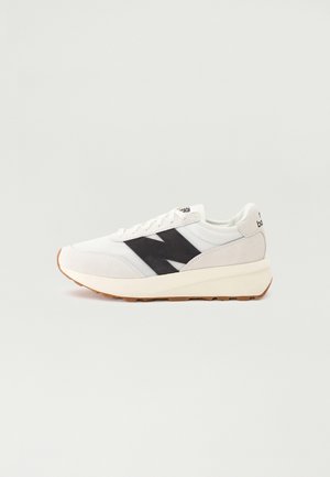 Biało-beżowy sneaker z czarnym logo "N" z boku, białymi sznurówkami oraz gumową podeszwą na jednolitym jasnym tle.