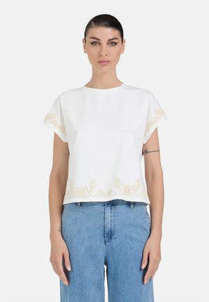 Donna con capelli scuri indossa una maglietta bianca a maniche corte con ricamo floreale e jeans blu a vita alta, in piedi davanti a uno sfondo semplice.