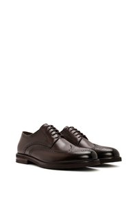 Une paire de chaussures habillées en cuir brogue marron foncé avec perforations en pointe et laçage fermé sur un fond blanc.