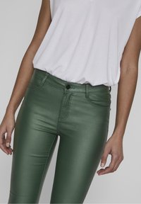 Pantalones vaqueros verdes, brillantes y ajustados con un acabado elegante, que cuentan con un cierre de botón en la parte delantera, lazos para cinturón y bolsillos frontales. Combinando con una blusa blanca.