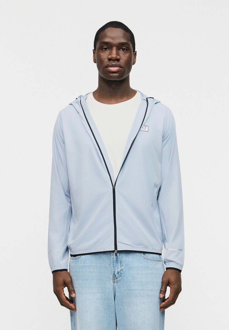 Νέος άνδρας με ανοιχτό μπλε φούτερ με φερμουάρ πάνω από λευκό πουκάμισο και ανοιχτό μπλε τζιν, στέκεται μπροστά σε απλό λευκό φόντο.