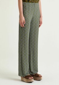 Pantaloni a gamba larga con motivo verde presentano un disegno geometrico in viola e bianco. Il tessuto sembra liscio, abbinato a sandali con zeppa marroni.