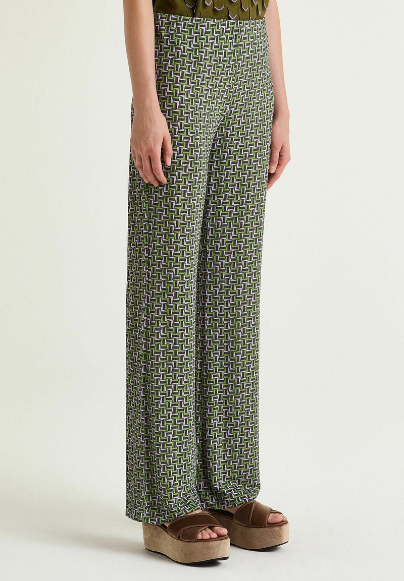 Pantaloni a gamba larga con motivo verde presentano un disegno geometrico in viola e bianco. Il tessuto sembra liscio, abbinato a sandali con zeppa marroni.