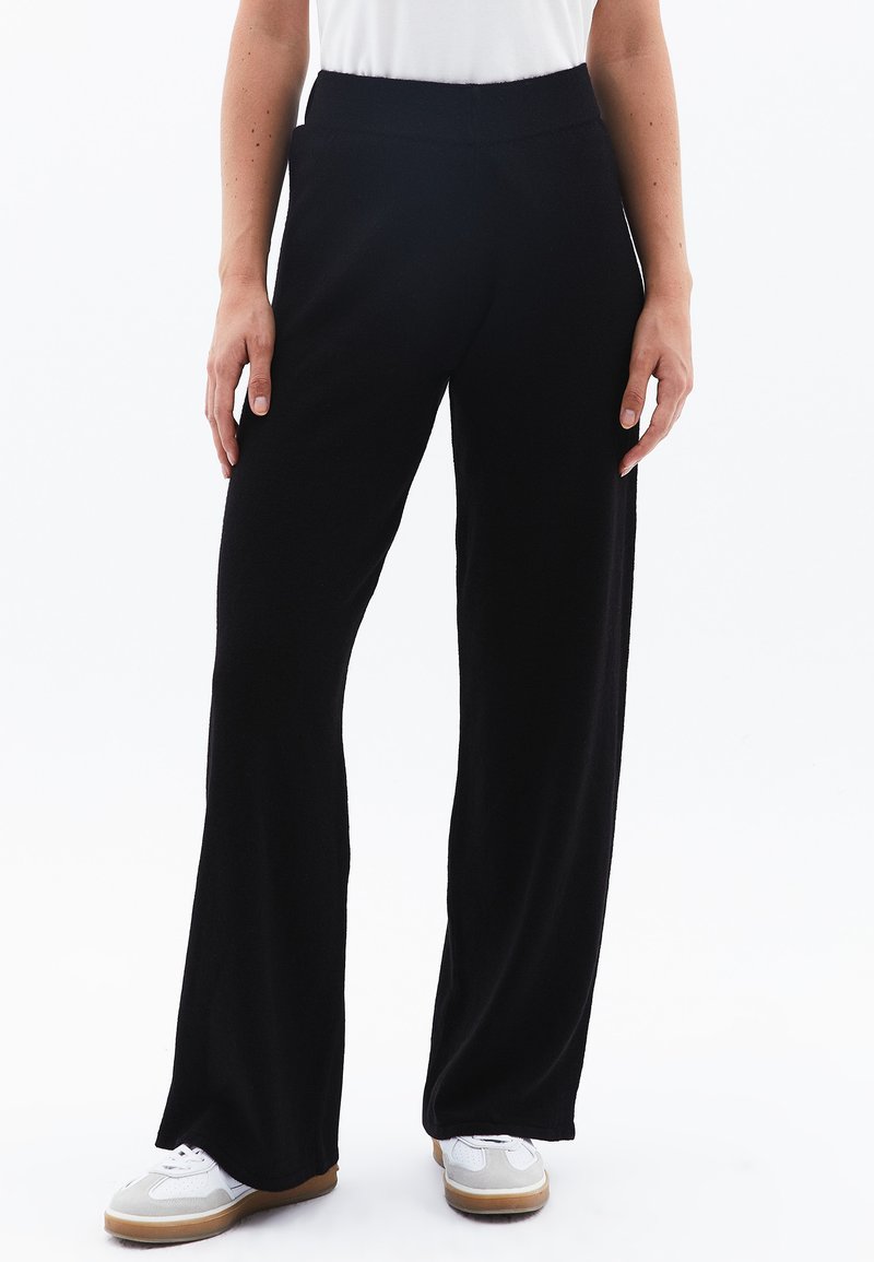 Pantaloni neri a gamba larga realizzati in tessuto morbido, con una cintura elasticizzata. Abbinati a una camicia bianca e scarpe sportive. Texture liscia, senza motivi.
