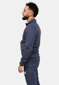 Tuta blu navy realizzato in tessuto morbido. Giacca con zip completa, colletto alto e tasche con zip, abbinata a pantaloni con vita elasticizzata.