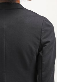 Musta bleiseri, jossa on vartalonmyötäinen leikkaus, sileä pinta ja terävät kaulukset. Takasauma kulkee pystysuoraan, ja hihat ovat mittatilaustyönä ilman koristuksia.