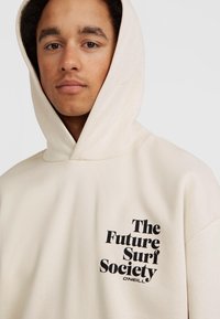 Felpa con cappuccio beige, dotata di tasca a marsupio frontale. Presenta la scritta nera stampata "The Future Surf Society" sul lato sinistro. Tessuto dalla texture morbida.
