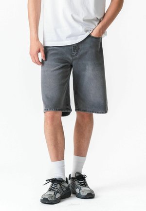 Man draagt verweerde grijze spijkershorts, witte sokken, zwart-grijze sneakers en een wit T-shirt, staat met één hand in zijn zak.