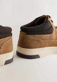 Brązowe zamszowe buty z czarnym materiałowym kołnierzem, ukazujące widoki z boku i z tyłu, podkreślające sznurówki i grube podeszwy na jasnym tle.