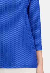 Blaues strukturiertes Oberteil mit einem Karomuster, lange Ärmel und abgerundetem Saum. Das Material wirkt leicht und dehnbar.