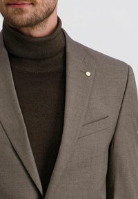 Behaarter Mann, der einen braunen Rollkragenpullover und ein taupefarbenes Sakko mit einem kleinen runden Anstecker am Revers trägt, vor einem schlichten Hintergrund.