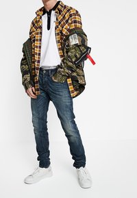 Man i lager av rutiga och kamouflagejackor över vit polo, blå jeans och vita sneakers, stående med en hand i fickan.