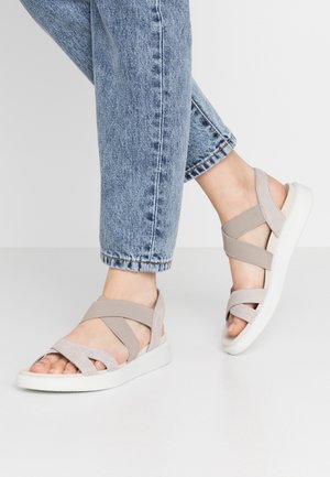 Stopy w beżowych sandałach na krzyż z białymi podeszwami, połączone z jasnoniebieskimi podwiniętymi dżinsami na gładkiej białej podłodze.