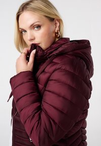 Femme aux cheveux blonds portant une veste matelassée bordeaux avec capuche, tenant le col près de sa bouche, regardant l'objectif.