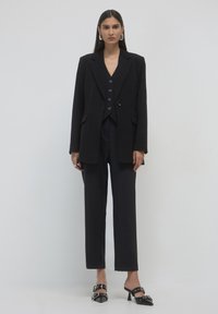 Black tailored blazer with matching high-waisted trousers, layered over a black waistcoat featuring buttons. Styled with black heeled mules.