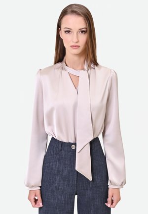 Donna con capelli castani lisci che indossa una camicetta rosa chiaro in raso con fiocco al collo e pantaloni grigio scuro a vita alta.