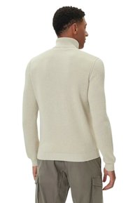 Maglione lavorato a maglia beige chiaro con collo alto, polsini a costine e un motivo testurizzato. Design aderente con maniche lunghe, indossato sopra pantaloni verdi.
