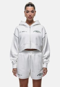 Witte cropped hoodie met een ritssluiting aan de voorkant en shorts met trekkoord. Kenmerkt zich door tekst in groen en grijs, gemaakt van zachte, gewatteerde stof.
