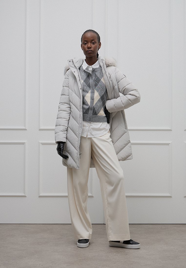 Jasnoszary płaszcz puchowy z kapturem, pikowany wzór; noszony na szarym swetrze w romby, białej koszuli i kremowych spodniach z szerokimi nogawkami, w połączeniu z czarnymi rękawiczkami.