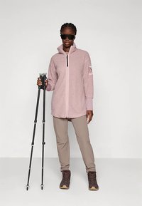 Kobieta w okularach przeciwsłonecznych, ubrana w różową polarową kurtkę, beżowe spodnie i buty trekkingowe, trzymająca dwa czarne kije trekkingowe.