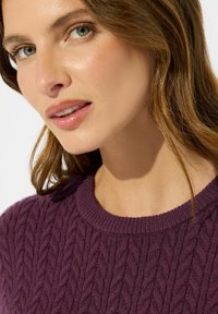 Maglione viola a treccia, con collo arrotondato, caratterizzato da dettagli di texture e una finitura morbida. Primo piano del collo e dei lineamenti del viso.
