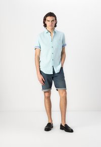 Himmelblaues Kurzarmhemd mit Kragen und Fronttasche, kombiniert mit dunklen Jeansshorts. Das Modell trägt schwarze Slipper.