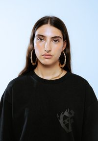 Zwart oversized t-shirt met een klein vlamlogo aan de linkerkant, gecombineerd met grote gouden oorringen en een gouden ketting.