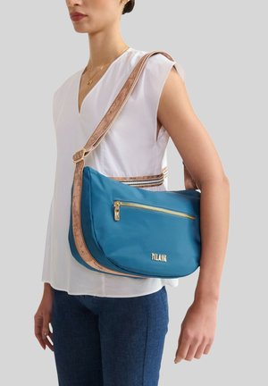 Donna che indossa una blusa bianca senza maniche e jeans blu, con una borsa a tracolla blu con tracolla beige e cerniera dorata.