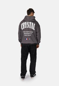 Felpa grigia con cappuccio e grande scritta bianca "CRYSTAL" sulla schiena. Abbinata a pantaloni neri e sneaker. Presenta un dettaglio della bandiera francese.