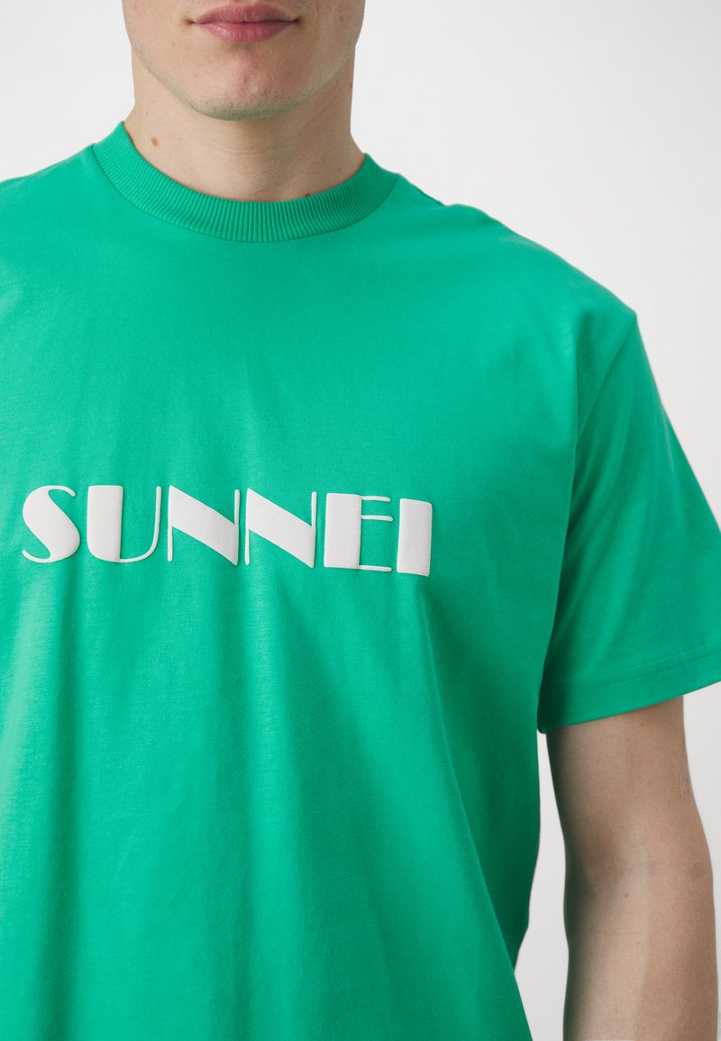 T-shirt verde in cotone con scollo a girocollo, caratterizzato da un logo stampato bianco "SUNNEI" di spessore variabile. Tessuto morbido, vestibilità comoda.