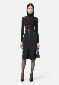 Robe noire à la coupe ajustée, avec une fermeture éclair à l'avant, une large ceinture et un haut en dentelle à manches longues sur un soutien-gorge. Des talons noirs et un sac à main complètent le look.