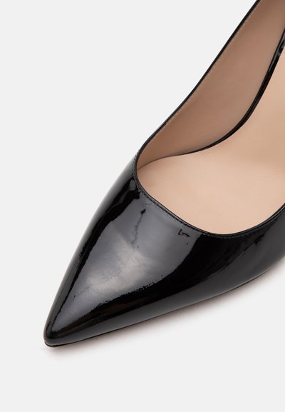 Chaussure à talon haut en cuir verni noir à bout pointu avec doublure intérieure beige sur fond blanc.