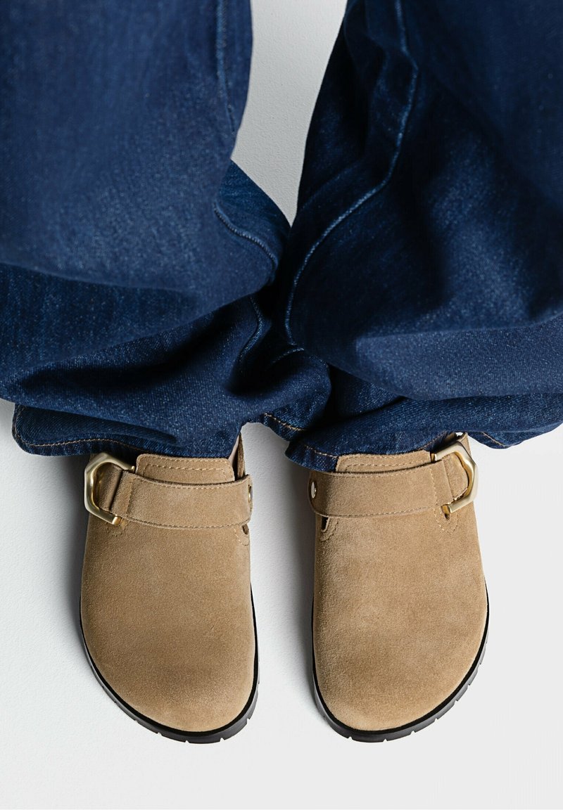Pieds portant des chaussures en daim beige à enfiler avec des boucles dorées, associées à un jean en denim bleu sur un sol blanc.