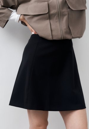 Άτομο που φοράει μαύρη φούστα τύπου A-line και μπεζ κοντό σακάκι με μεγάλες μπροστινές τσέπες, το χέρι ακουμπισμένο στη μέση και φωτεινό φόντο.