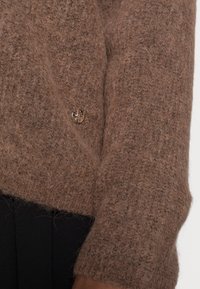 Maglione a maglia marrone con una finitura morbida e testurizzata. Presenta un bottone dorato e una vestibilità ampia, abbinato a un tessuto nero sottostante.