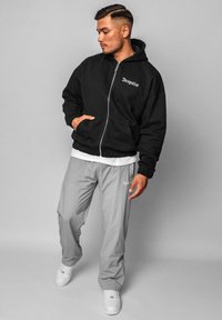 Sweat à capuche noir avec fermeture éclair, poches avant et lettres blanches. Associé à un pantalon gris clair ample avec un logo. Des baskets blanches complètent le look.