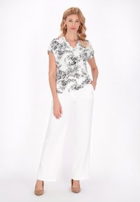 Blusa floreale bianca con motivi neri, maniche corte e scollo a V, abbinata a pantaloni bianchi a palazzo e tacchi beige.