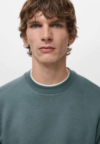 Maglione grigio-verde con scollo classico a girocollo, realizzato in tessuto morbido e strutturato, indossato sopra una camicia bianca, su un modello con capelli di media lunghezza.