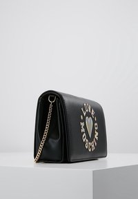 Svart läderhandväska med en strukturerad form, utrustad med ett guldchain-rem och en iriserande hjärt- och bokstavsdetalj på framsidan.