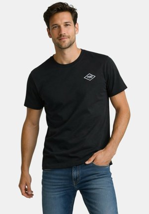 Homme portant un t-shirt noir à manches courtes avec un petit logo blanc sur la poitrine et un jean bleu, debout avec une main dans la poche.