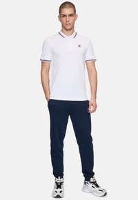 Vit polo-shirt med lila och röda ränder på kragen, ihop med marinblå joggingbyxor och vita sneakers med svarta accenter.