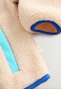 Beżowa kurtka polarkowa z niebieskimi akcentami, z turkusowym panelem, miękką fakturą i małą metką "little bird" blisko krawędzi.