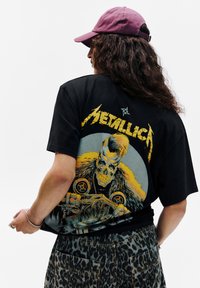 Černé oversized tričko s barevným potiskem na zádech, představujícím lebku a žluté akcenty. Slučuje se s kalhotami s leopardím potiskem.